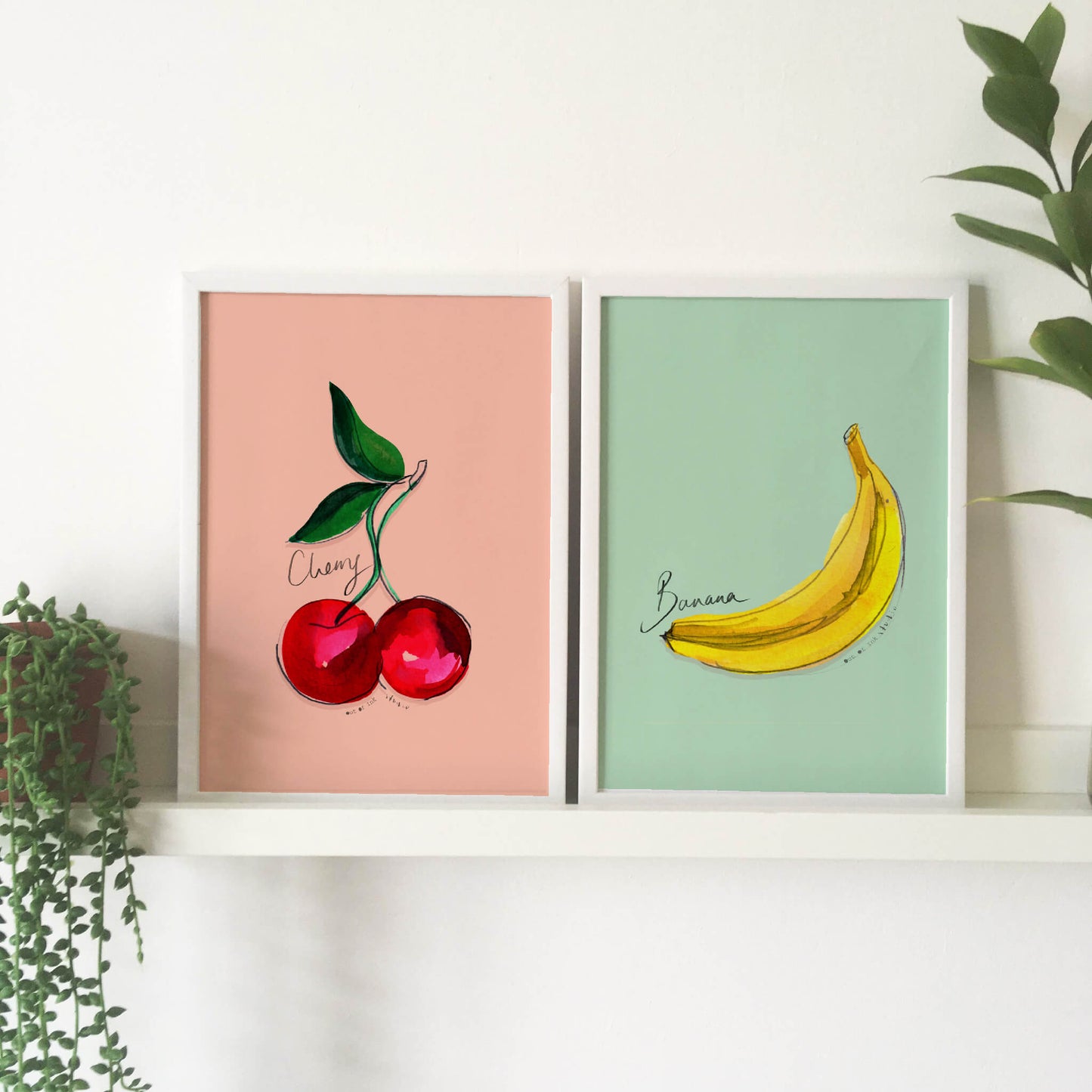 Two prints side by side. One is a hand-painted cherry on a pink background. The other is a hand-painted banana on a green background with text ‘banana’ hand written on it.