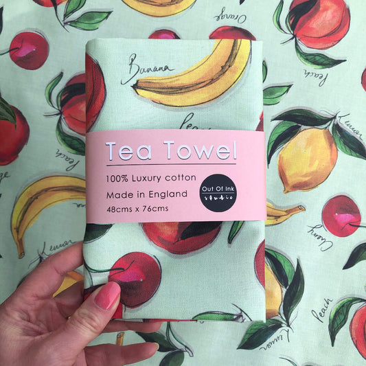 A tea towel featuring hand-painted fruit, banana, lemons, peaches, oranges and cherries.