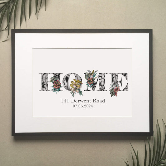 A print of the word 'HOME' filled with tropical imagery. Underneath the print is personalised with address and moving in date. hung on wall in black frame above a green sofa.